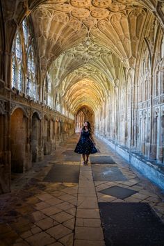 a woman in a dress walking down a long hallway with arches and stone flooring