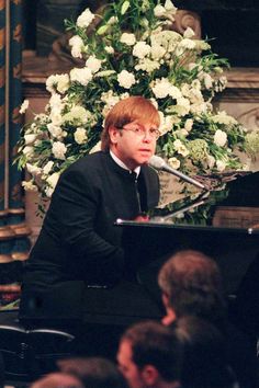 a man sitting at a piano in front of a microphone with flowers on the wall behind him