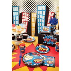 a party table set up with comic themed plates and cups, napkins and cupcakes