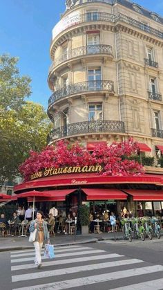 people walking across the street in front of a building with red flowers on it's roof