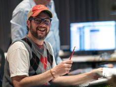 a man sitting in front of a computer desk holding a pen and smiling at the camera