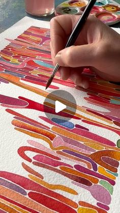 a person is drawing on paper with a pen and watercolors in front of them