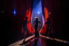 three people standing in front of red curtains on stage with spotlights shining through them