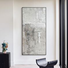 an abstract painting hangs on the wall next to a black chair and vase in a white room