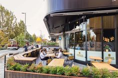 people sitting at wooden tables in front of a building with large windows and plants growing on the outside