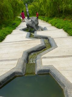 two people are walking on a path near a small pond and some rocks with water running through it