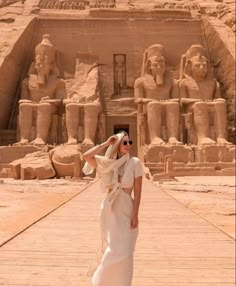 a woman standing in front of an ancient building with statues on the walls and behind her she is wearing a white dress