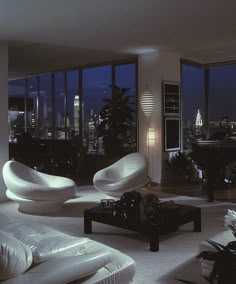 a living room filled with white furniture next to large windows at night time in front of the cityscape