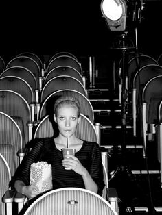 a black and white photo of a woman sitting in a chair holding a drink while looking at the camera