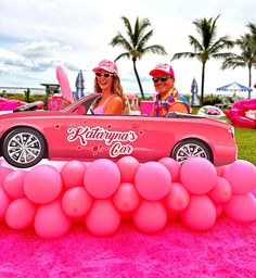 two women sitting in a pink car with balloons on the ground and palm trees behind them