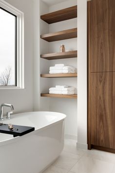 a white bath tub sitting under a window next to a wooden shelf filled with towels