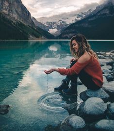 a woman sitting on rocks in the water with mountains in the background and clouds in the sky