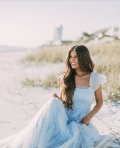 a beautiful woman sitting on top of a sandy beach next to the ocean wearing a blue dress