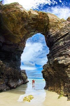 two people are standing in the water under an arch at the beach with rocks on either side