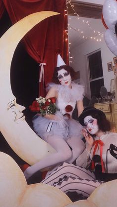 two women dressed in costumes sitting on top of moon shaped objects and one is holding flowers