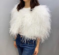 a woman standing in front of a gray wall wearing a white feathered jacket and jeans