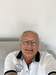 an older man wearing glasses sitting on a couch with his arms crossed and looking at the camera