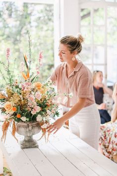 a woman arranging flowers in a vase on top of a white table with people sitting around