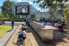 people sitting on benches in front of a building with a large screen above it that reads,