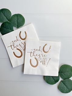 two white napkins with gold foil lettering on them and green leaves surrounding the edges