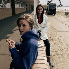 two women are sitting on a bench near the beach and one is looking at the camera