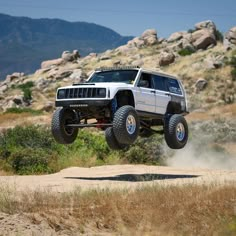 a white jeep driving through the air on top of a dirt road with rocks in the background