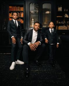 three young men in suits and ties sitting on a couch with bookshelves behind them