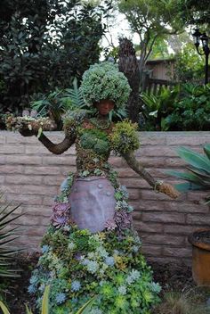 a woman made out of plants standing next to a brick wall