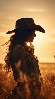 a woman with long hair wearing a cowboy hat standing in a wheat field at sunset