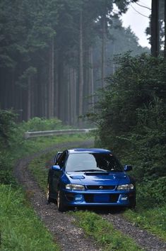 a blue car parked on the side of a dirt road in front of tall trees