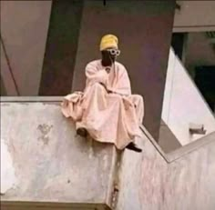 a man sitting on the side of a building wearing a yellow hat and glasses with his hands in his pockets