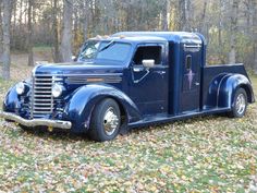 an old blue truck is parked in the grass near trees and fallen leaves on the ground