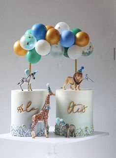 two cakes decorated with animals and balloons on top of a white cake stand in front of a gray wall