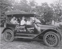 black and white photograph of people in an old car with advertising on the front window