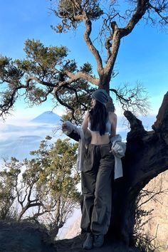 a woman standing on top of a mountain next to a tree and talking on a cell phone