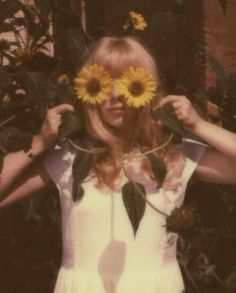 a woman with sunflowers on her face standing in front of some leaves and flowers