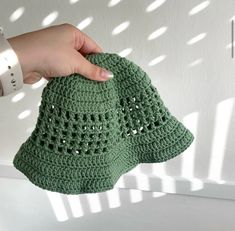 a hand holding a green crocheted hat on top of a white surface with sunlight coming through the window