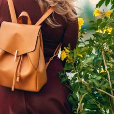 a woman wearing a tan leather backpack standing in front of yellow flowers