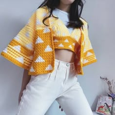 a woman wearing white pants and a yellow knitted jacket with triangles on it, standing in front of a wall