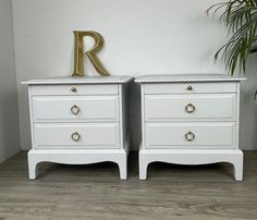 two white nightstands with gold handles and letters on them in front of a potted plant