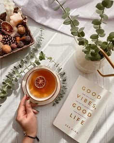 a person holding a cup of tea next to a book and potted plant on a table