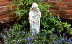 a statue in the middle of some blue flowers and plants next to a brick wall