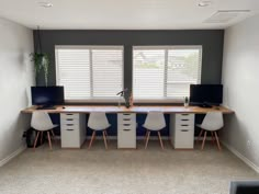 a desk with two computers on top of it in front of three white chairs and windows