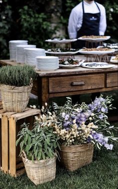 a table topped with lots of baskets filled with flowers next to a man in an apron