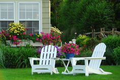 two white chairs sitting on top of a green grass covered lawn next to flowers and bushes