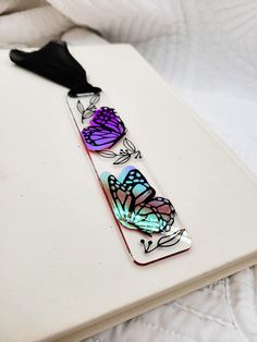 a bookmark with butterflies on it sitting on a white bed sheet, next to a black tassel