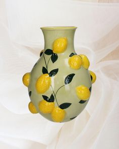 a green vase with lemons painted on the side and leaves in the middle, sitting on a white cloth