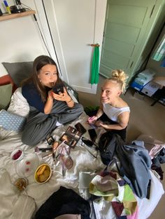 two young women sitting on a bed with clothes and other items scattered about the room