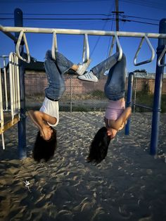 two girls hanging upside down on the playground equipment at an outdoor play area with sand and blue skies in the background