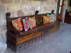 a wooden bench with four colorful pillows on it's back and two windows in the background